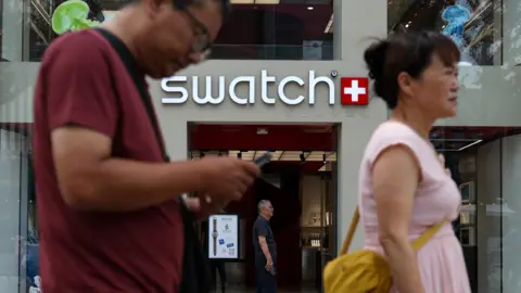 Reuters Leute gehen an einem Geschäft von Swiss Watchmaker Swatch in Peking, China, vorbei. Eine Frau und ein Mann sind im Vordergrund zu sehen. Er trägt ein kastanientiertes T-Shirt und sie trägt ein rosa Oberteil mit einer gelben Tasche. Das Swatch -Logo scheint hinter ihnen zu sehen, mit dem Eingang zum Laden unten