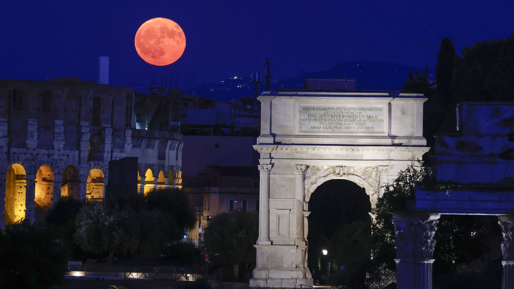 A reddish full moon can be seen rising against a night sky, with the ancient, curved outer wall of the colosseum in Rome positioned beneath, illuminated from within by a yellow light. A large arch is starkly illuminated to the right of the image, towering above nearby trees.