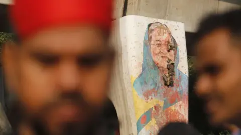 Nurphoto über Getty Images Ein Gemälde von Sheikh Hasina trägt einen blauen Schal und mit Bangladesch -Flaggen unter ihr auf einer Betonsäule scheint Löcher darin zu haben und ist mit Splatten aus roter Farbe bedeckt. Das verschwommene Gesicht eines Mannes in einem roten Hut ist im Vordergrund zu sehen