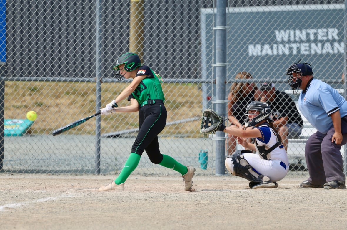 Ein Frauen -Softball -Teig für Frauen schwingt auf einem Spielfeld, während ein Fänger hinter sich hockt. Ein Schiedsrichter hockt hinter dem Fänger.