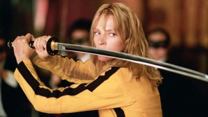 Eine Frau mit blonden Haaren, die eine gelbe und schwarze Jacke tragen, hält ein Samurai -Schwert mit beiden Händen, die nach vorne gerichtet sind. Der Hintergrund ist verschwommen, was auf eine Inneneinstellung mit anderen vorhandenen Zahlen hindeutet. Die Person erscheint fokussiert und bestimmt.