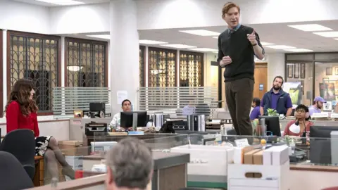 Peacock Domhnall Gleeson steht auf einem Tisch in einem Büro im Charakter als Ned Sampson