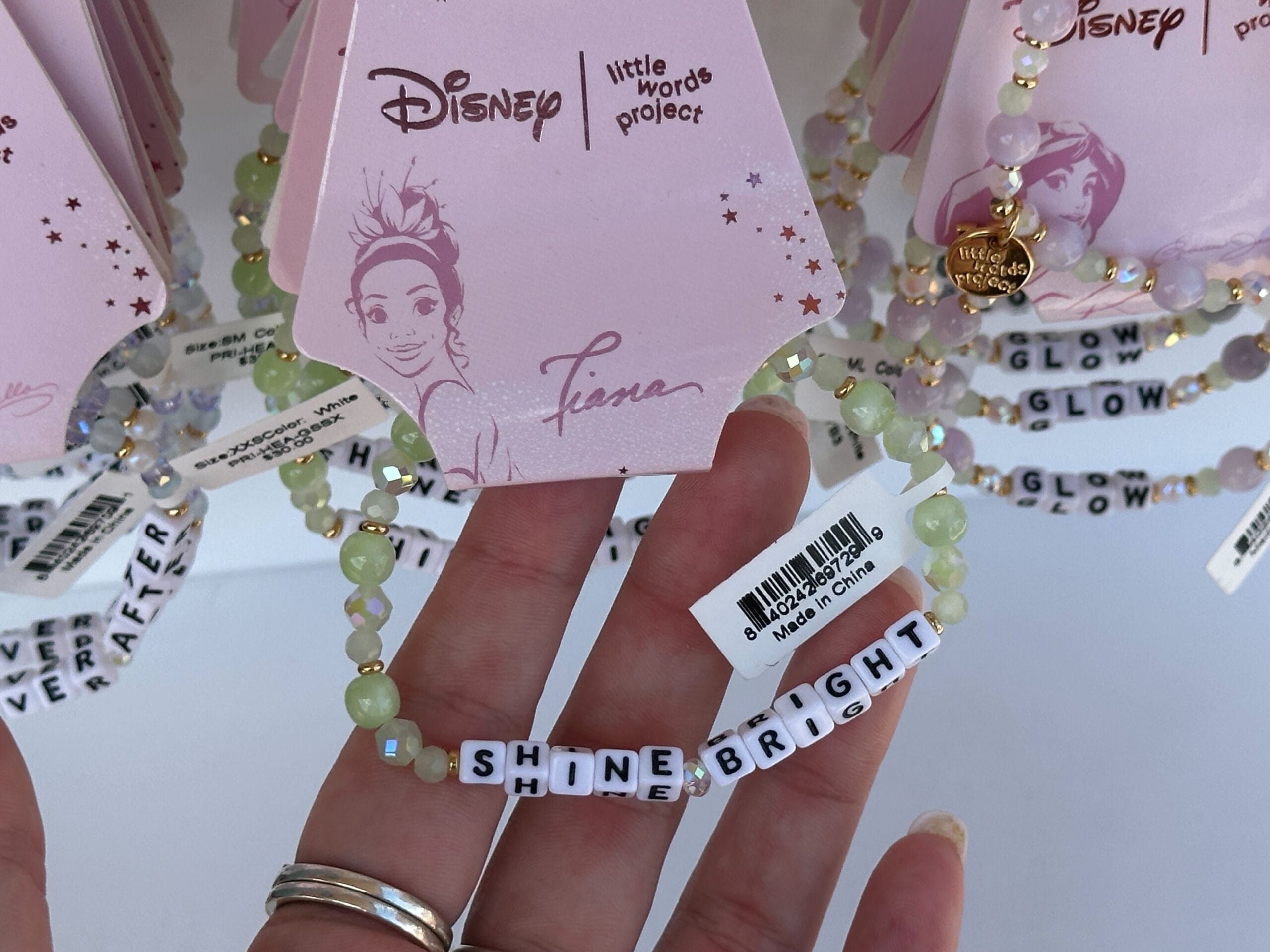 Eine Hand hält ein Perlenarmband mit Buchstabenperlen Rechtschreibung "Leuchtet hell" Unter Disney Princess Tiana Verpackung.