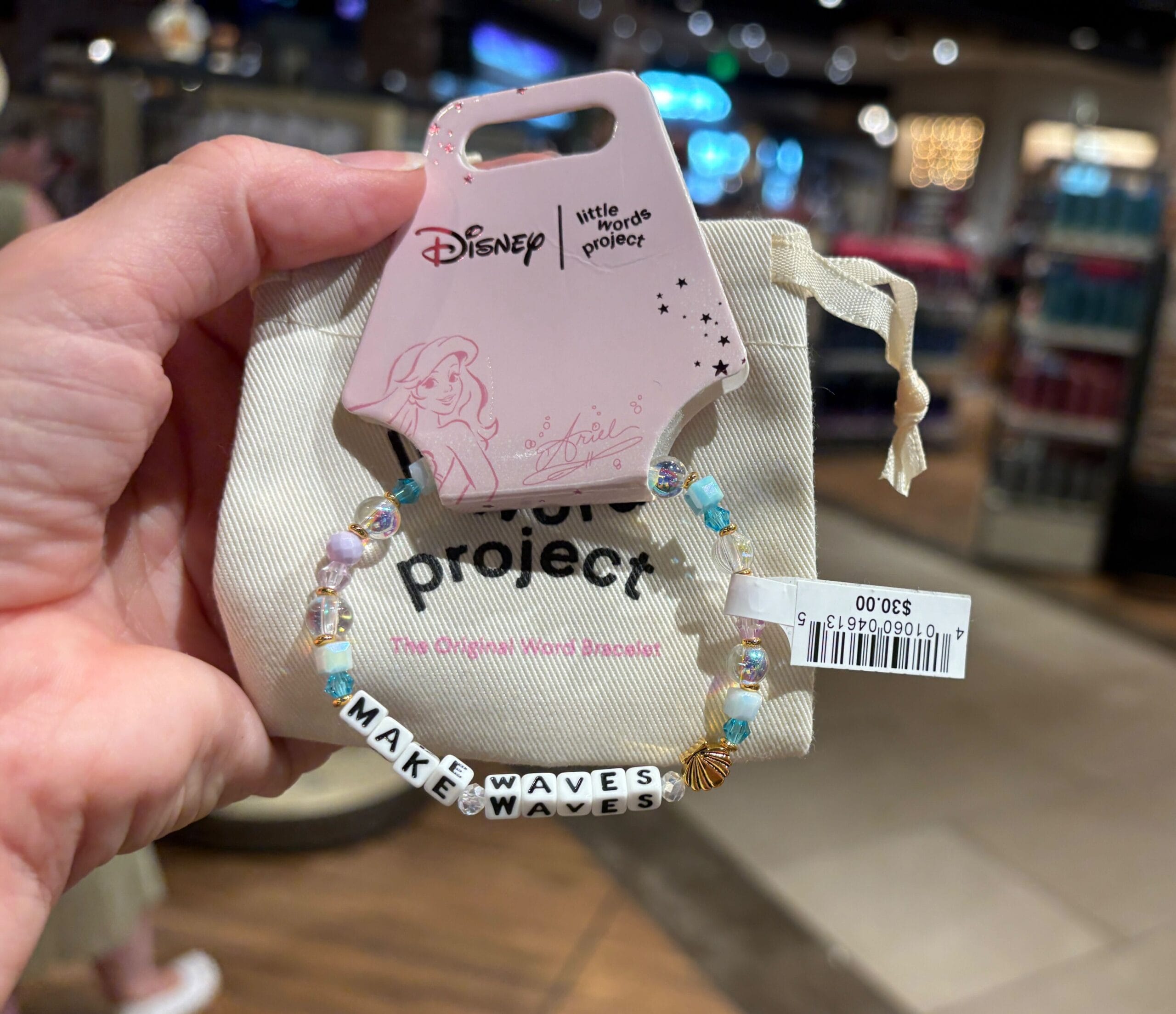 Eine Hand hält einen Disney "Wellen machen" Perlenarmband mit Ariel auf dem Etikett in einem Geschäft.