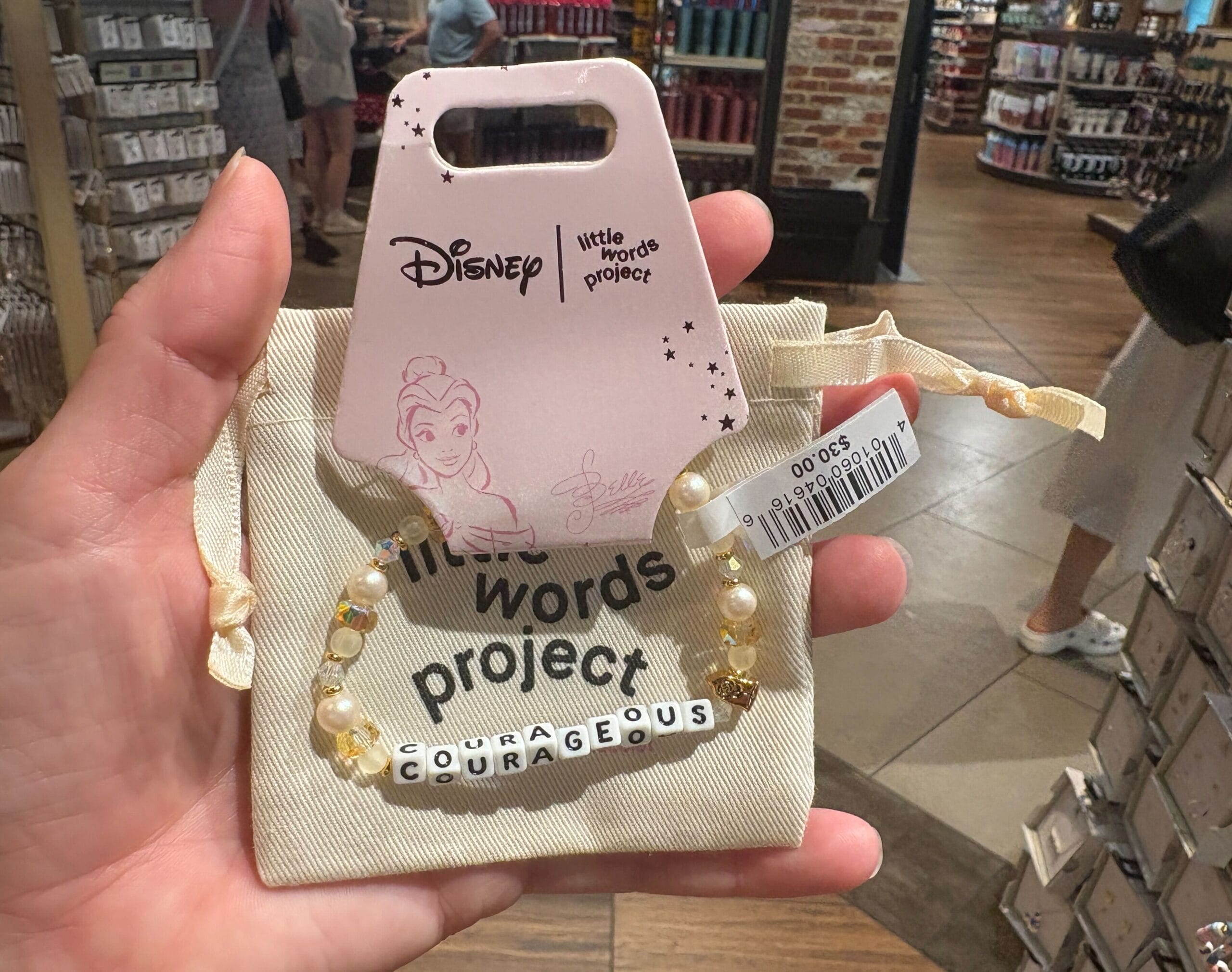 Eine Hand hält einen Disney "Mutig" Perlenarmband mit Belle auf Pink -Tag in einem Geschäft.
