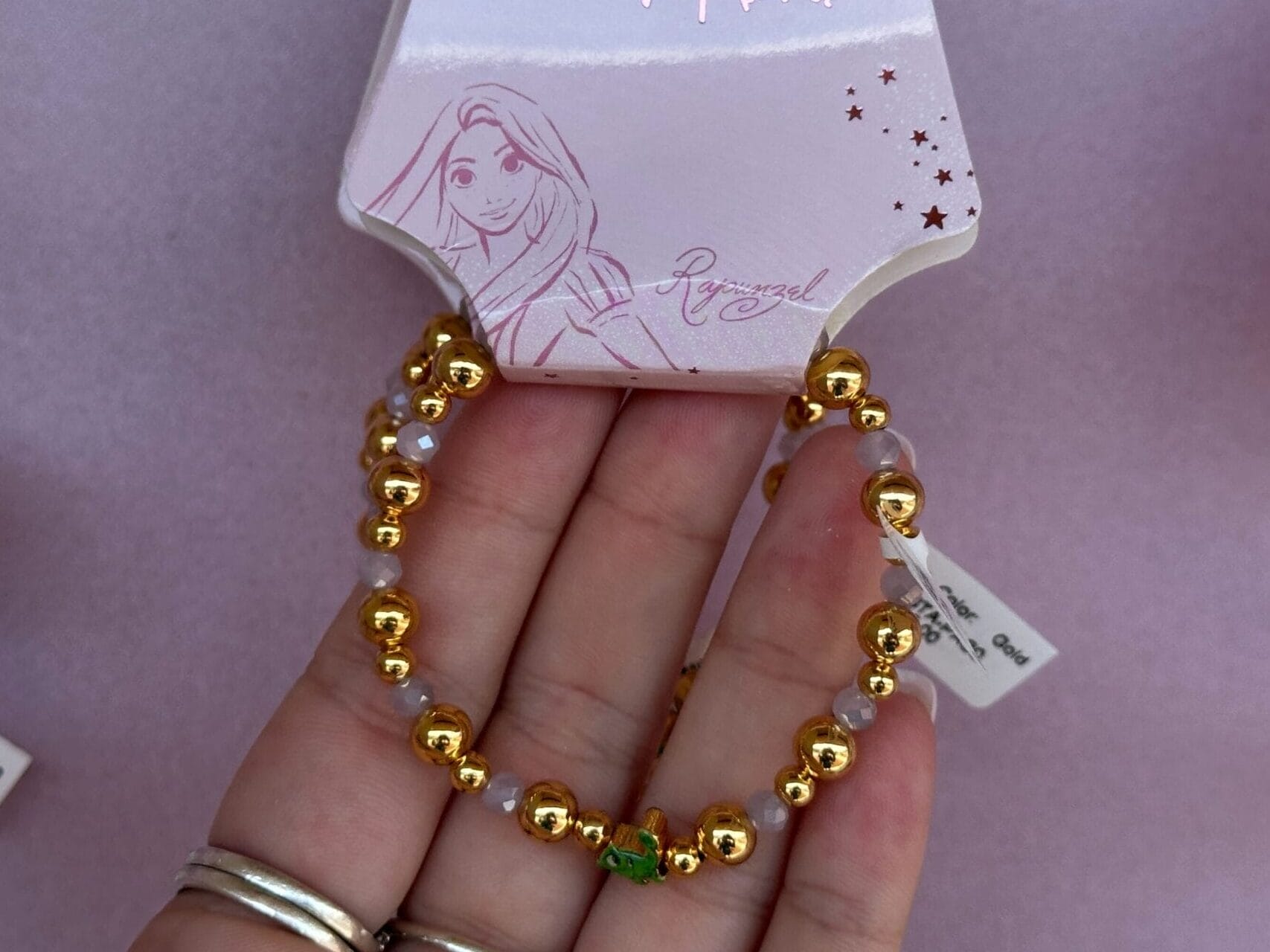 Hand hält ein Disney Rapunzel -Armband mit goldenen Perlen und einem grünen Akzent, der auf rosa Verpackungen angezeigt wird.