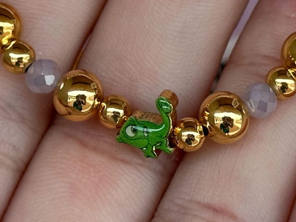 Hand hält goldenes Perlenarmband mit einem kleinen grünen Schlangenbrand und einigen facettierten blassen Perlen vor einem rosa Hintergrund.