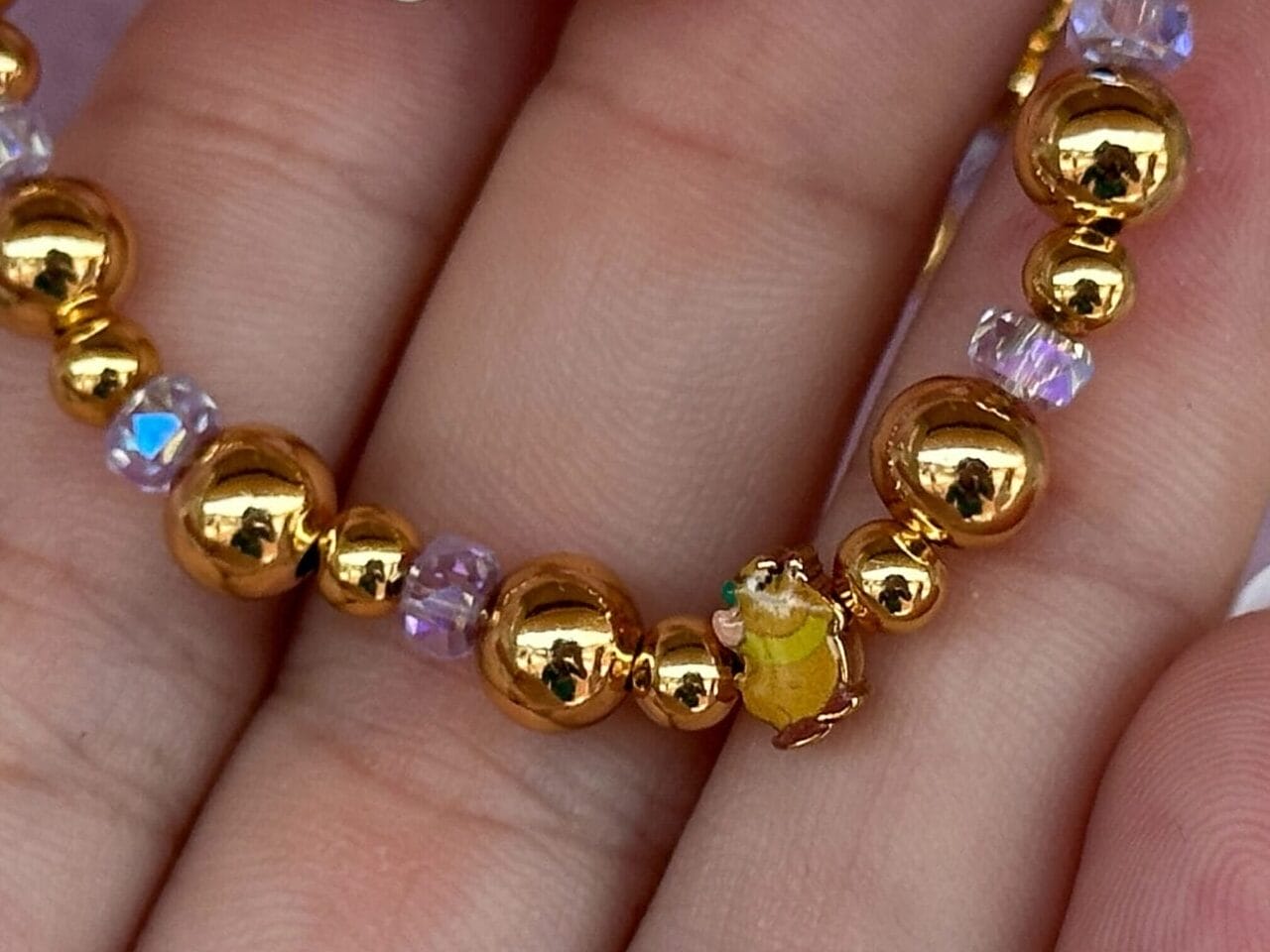 Eine Hand hält ein goldenes Perlenarmband mit lila Akzenten und einer gelben Perle vor einem rosa Hintergrund.