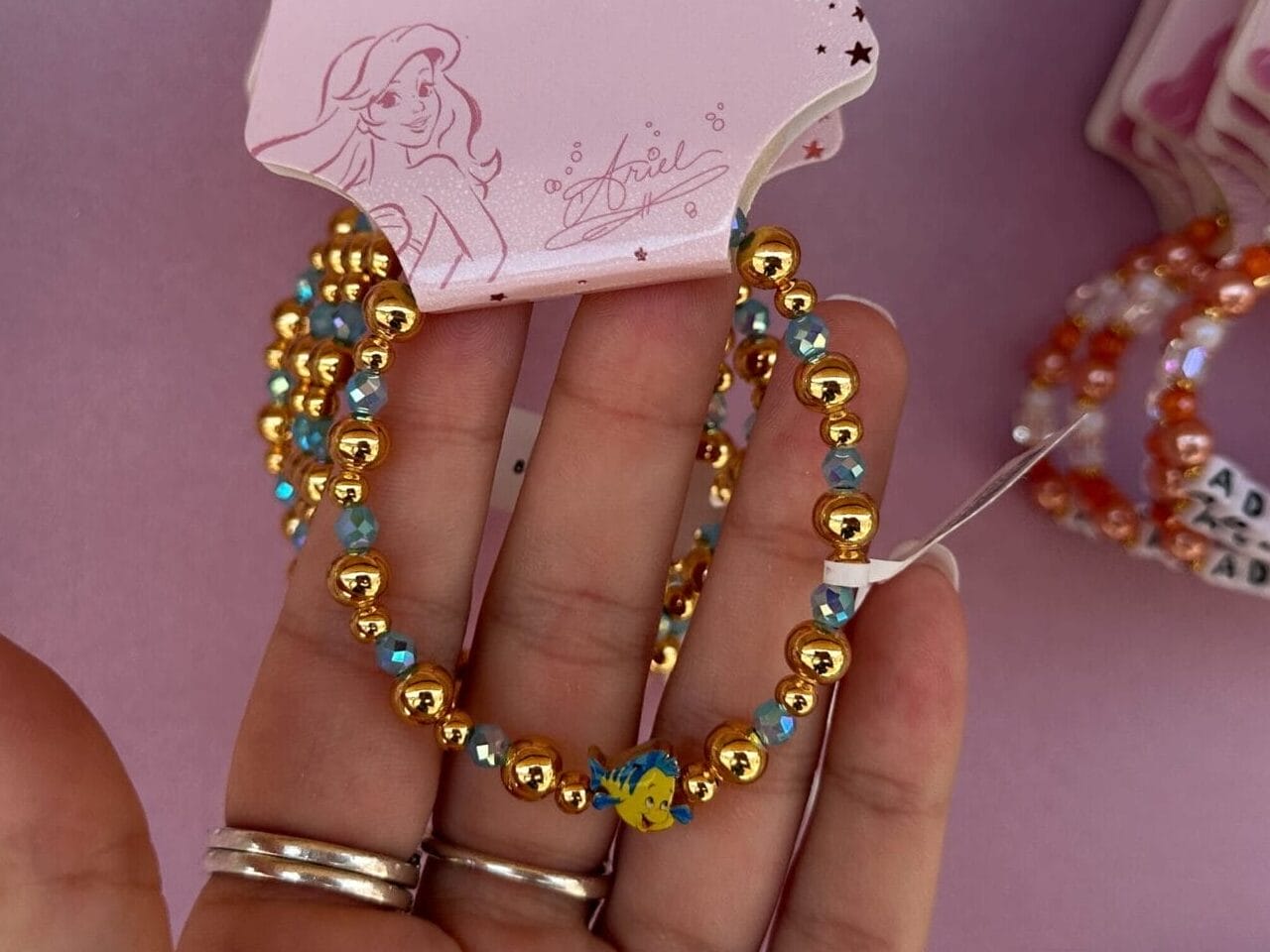Eine Hand hält Disney Princess Perlen Hoop -Ohrringe gegen eine Ausstellung ähnlicher Ohrringe auf einem rosa Hintergrund.