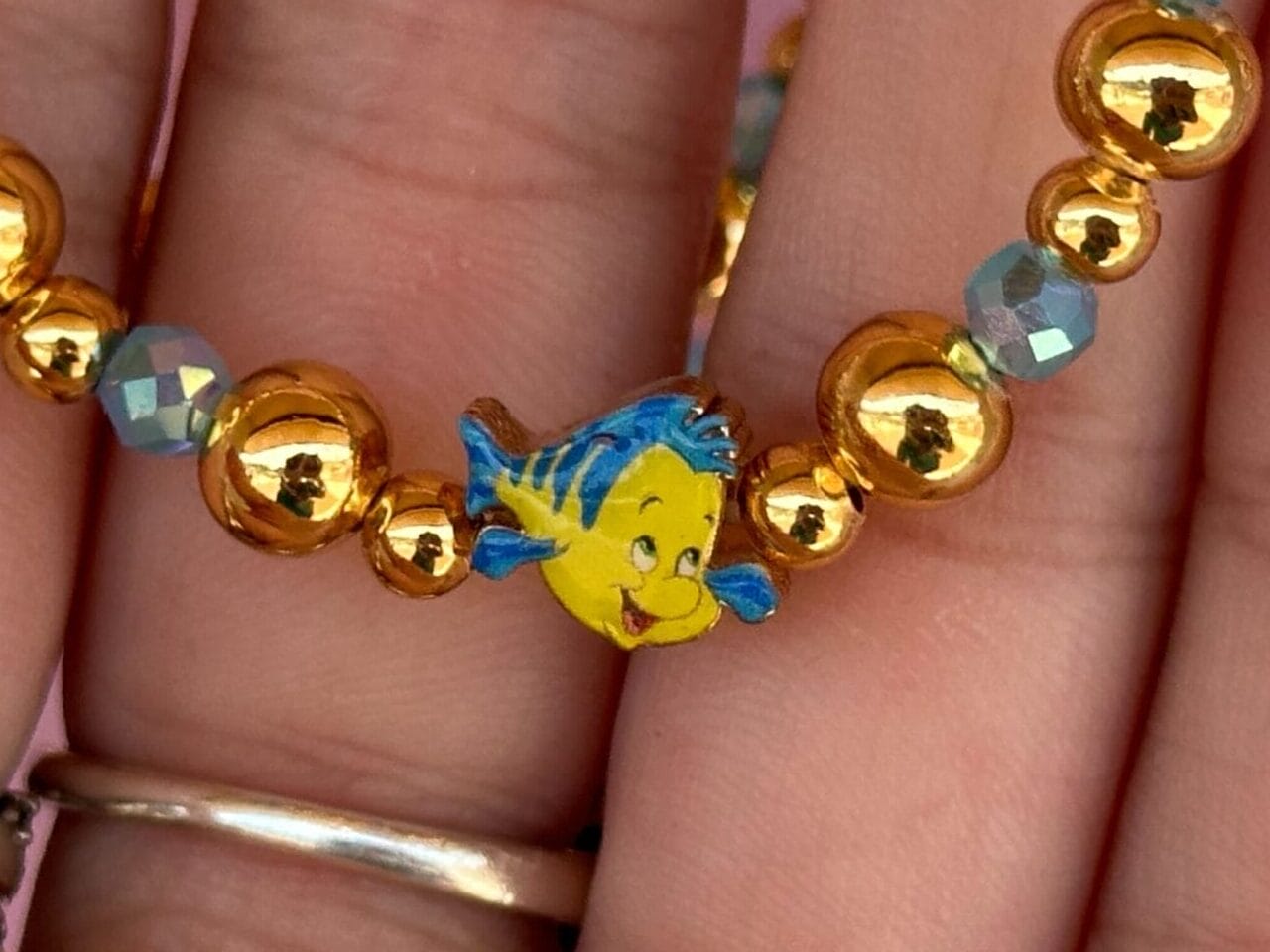 Eine Hand hält ein Perlenarmband mit goldenen Perlen und einem kleinen blauen und gelben Fischbrand.
