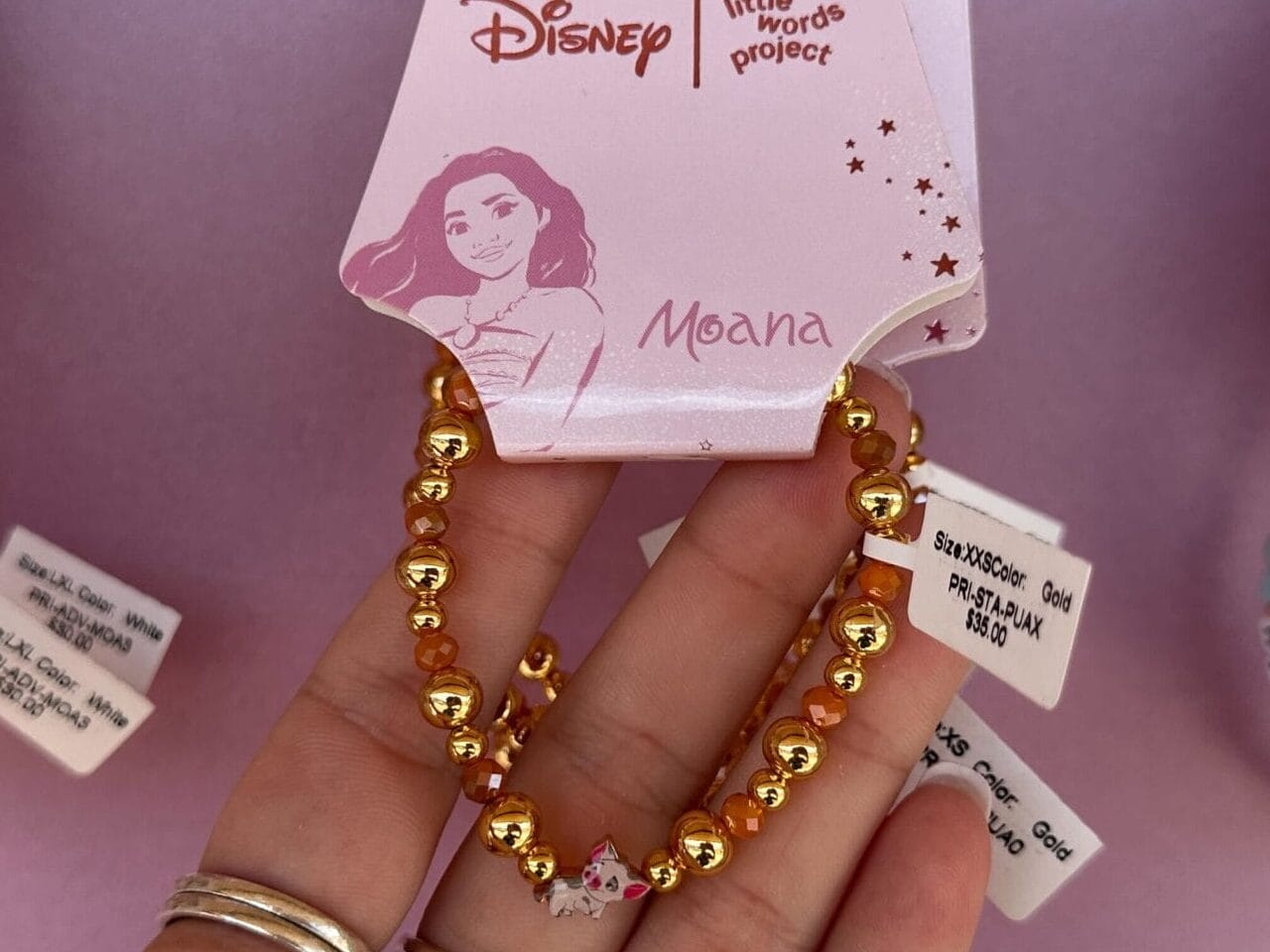 Hand hält ein Disney-Armband zum Thema Moana mit goldenen Perlen, die unter anderem auf einem rosa Hintergrund angezeigt werden.