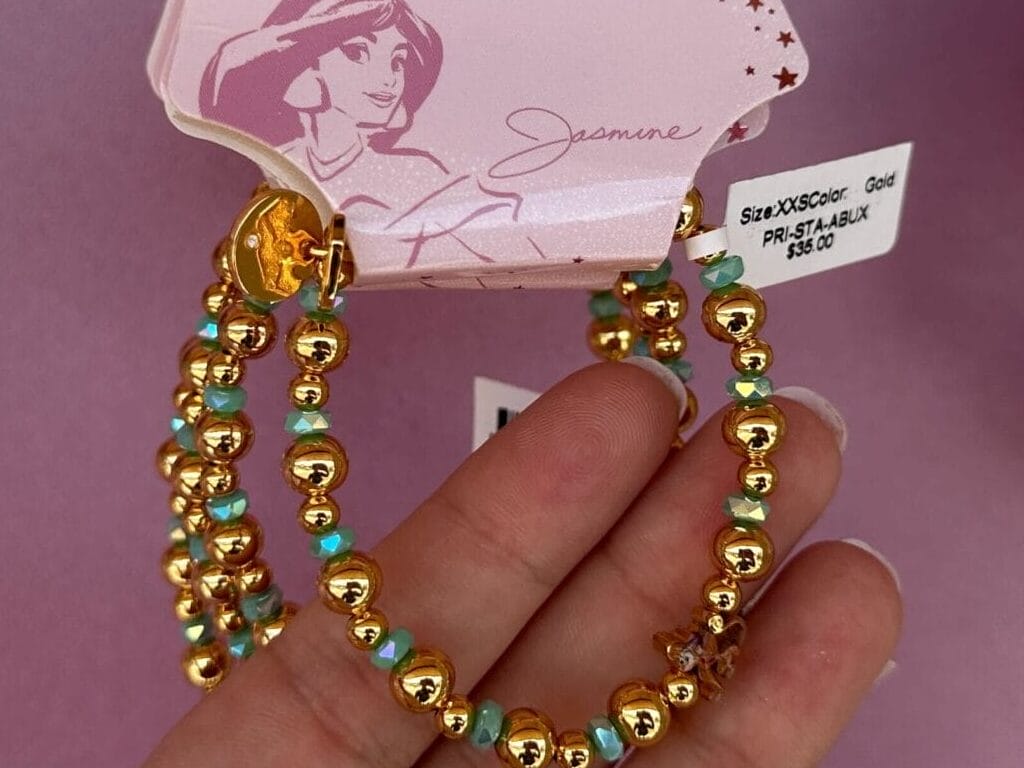 Eine Hand hält ein Disney -Prinzessin Jasmine Perlen -Armband -Set mit Gold- und türkisfarbenen Perlen vor einem rosa Hintergrund.