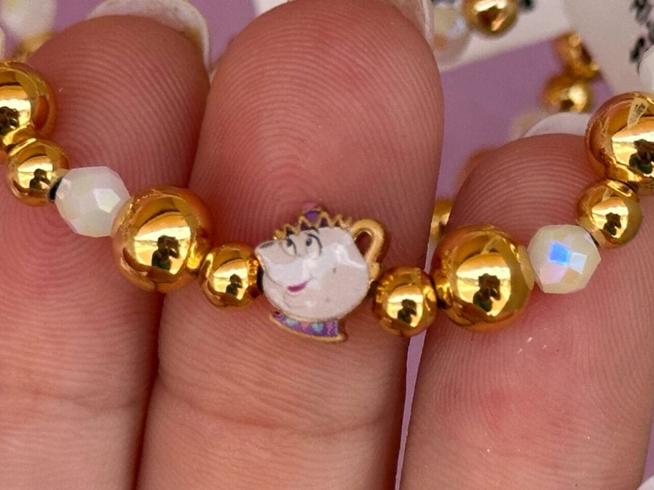 Hand hält ein goldenes und weißes Perlenarmband mit einem kleinen Chip, der Teetasse aus Schönheit und Biest.