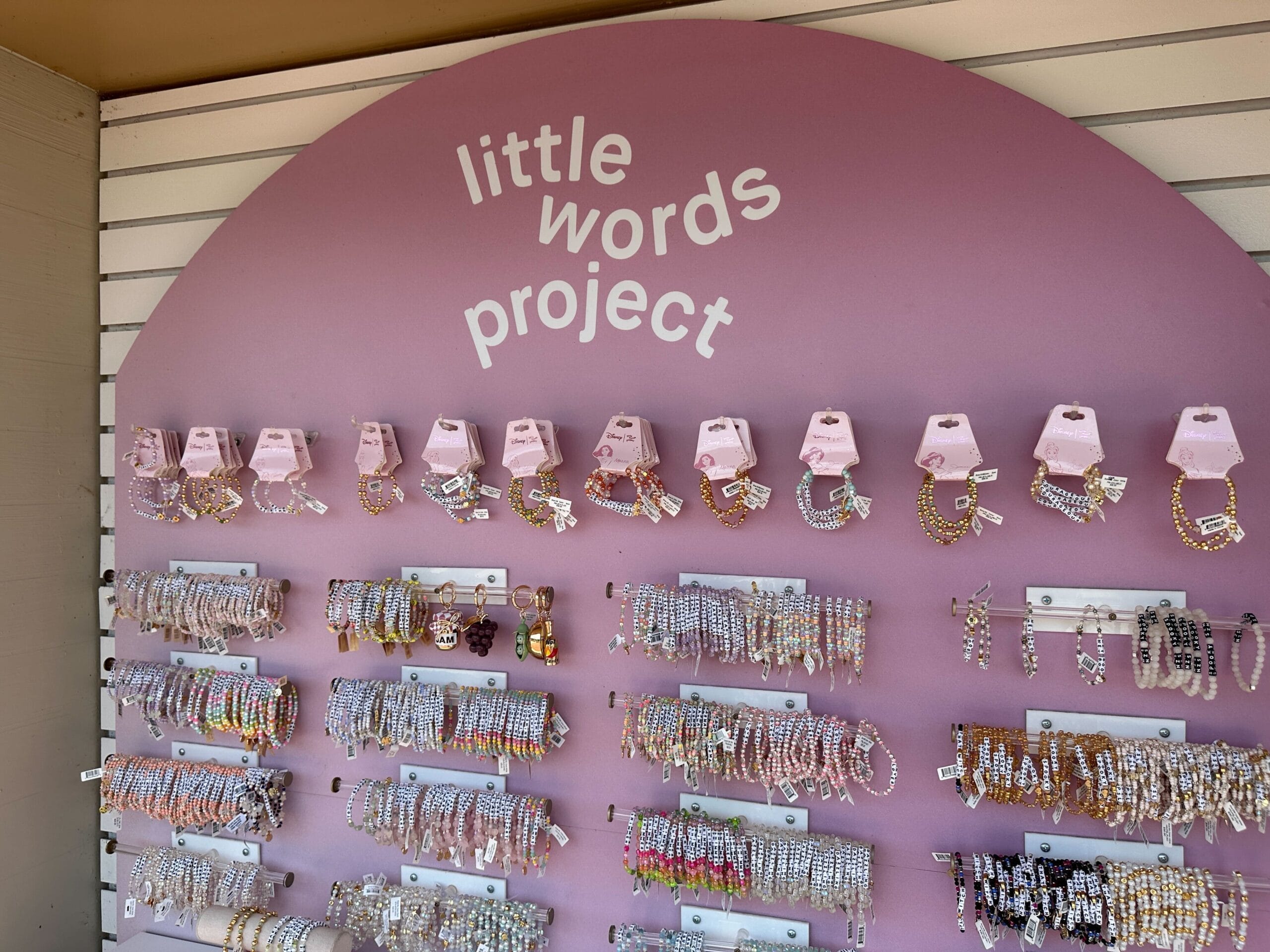 Eine Anzeige von farbenfrohen Perlenarmbändern und Schlüsselanhagen unter einer Schildlesung "Little Words Project" auf lila Hintergrund.
