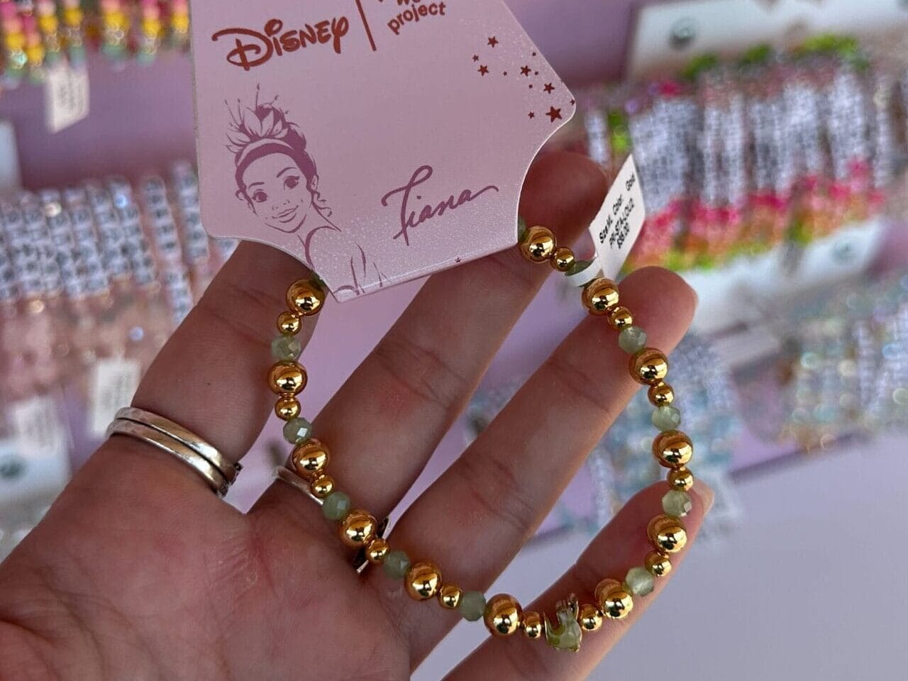 Eine Hand hält ein goldenes Brakband von Disney Princess Tiana vor einer Ausstellung farbenfroher Armbänder.